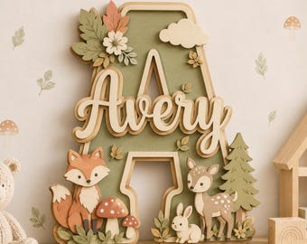May include: A wooden letter "A" with the name "Avery" in cursive, decorated with woodland creatures and foliage. The design includes a fox, deer, rabbit, mushrooms, and a tree, all in natural colors.