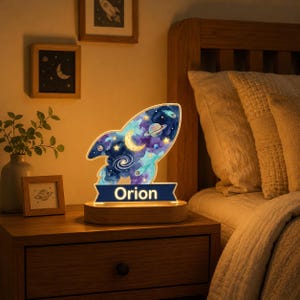 May include: A rocket-shaped LED night light featuring a colorful space-themed print with planets, stars, and a crescent moon. The base is made of light wood and includes a dark blue banner with the name Orion illuminated in white letters.