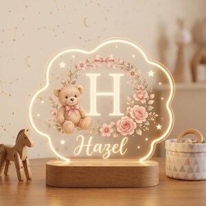 May include: A cloud-shaped LED night light with a wooden base featuring a light-up letter H, a teddy bear, and a floral wreath. The name Hazel is printed in script below the letter. The design includes small stars and a pink bow accent.