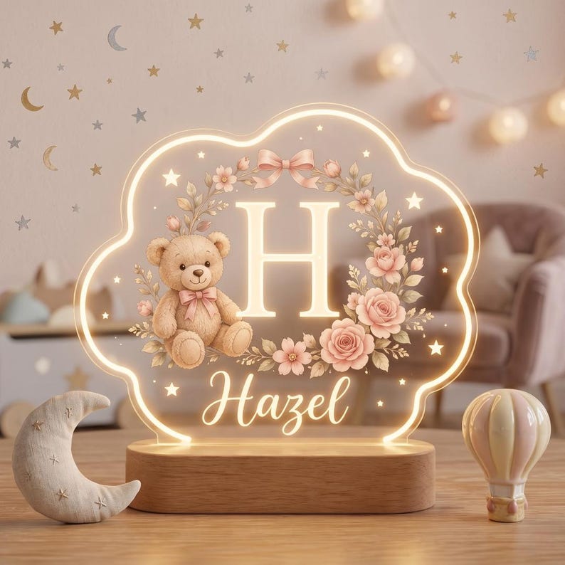 May include: A cloud-shaped LED night light with a wooden base, featuring a printed design of a teddy bear, a floral wreath, the letter H, and the name Hazel in script. The light glows with a warm yellow border and internal illumination.