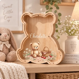 Può includere: Un salvadanaio in legno a forma di orso con un pannello frontale trasparente. L'interno presenta una grafica stampata di un orsacchiotto seduto su una tovaglia da picnic a quadretti rossi e bianchi con un cestino, fragole e margherite. Il nome Charlotte è stampato.