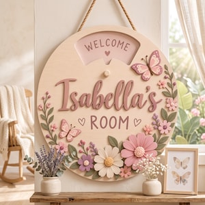 May include: A round wooden wall sign featuring the text "WELCOME" at the top and "Isabella's ROOM" in the center. The sign is decorated with three-dimensional pink and white flowers, green leaves, and two butterflies, all suspended by a rope.