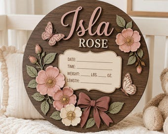 May include: A circular dark wood sign featuring the name Isla Rose in raised lettering. It includes a central plaque with fields for date, time, weight in lbs and oz, and length. Decorated with 3D pink flowers, green leaves, butterflies, and a bow.