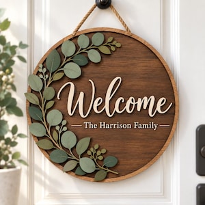 May include: A circular wooden sign featuring a dark brown wood grain finish, suspended by a rope. The sign displays the word "Welcome" in large, light-colored script lettering, with "The Harrison Family" printed below, accented by green eucalyptus leaves.