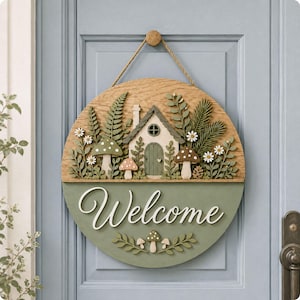 May include: A round wooden door sign featuring a layered design with a cottage, ferns, mushrooms, and white daisies. The bottom half is painted sage green with the word Welcome in white script, accented by small mushrooms and leafy sprigs.