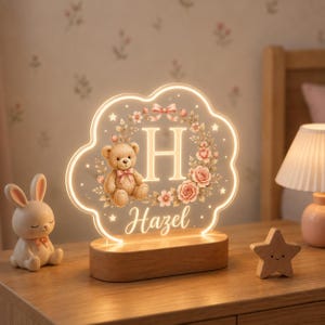 May include: A cloud-shaped LED night light with a wooden base, featuring a central letter H, a teddy bear, and a floral wreath design. The name Hazel is printed in script below the letter, and the entire acrylic panel glows with warm light.