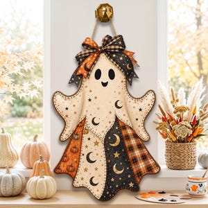 May include: A hanging ghost-shaped wall decoration featuring a cream-coloured body with black crescent moons and stars. The lower section includes panels of orange pumpkins, black moons, and orange plaid. A large plaid bow sits at the top.