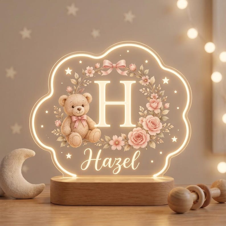 May include: A cloud-shaped LED night light featuring a light-up acrylic panel with a tan teddy bear, a floral wreath, the letter H, and the name Hazel in script. The panel is mounted on a rectangular wooden base with a warm glowing light border.
