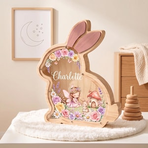 Könnte beinhalten: Eine hölzerne Spardose in Hasenform mit rosa bemalten Ohren und einer durchsichtigen Frontplatte. Die Platte zeigt den Namen Charlotte in weißer Schreibschrift, umgeben von Illustrationen einer Fee, eines kleinen Hasen, eines Pilzhauses und Blumen.