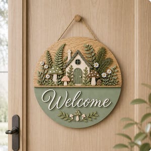May include: A round wooden door sign featuring a layered 3D design of a small cottage surrounded by ferns, pine branches, and mushrooms. The lower section is painted sage green with the white script text Welcome and small mushroom accents.