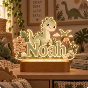 Puede incluir: Un letrero de acr&iacute;lico iluminado con un dinosaurio de dibujos animados, un volc&aacute;n y el nombre Noah en letras grandes. Un poste de madera dice Little Explorer Big Adventures. La pieza est&aacute; montada sobre una base de madera ovalada y brilla.
