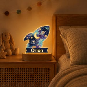 May include: A rocket-shaped LED night light featuring a watercolor space design with a crescent moon, planets, and stars. The base is made of light wood and includes a dark blue banner at the bottom with the name Orion printed in white text.