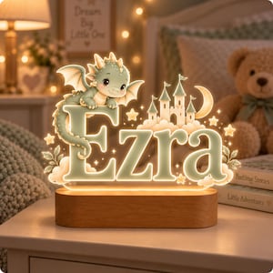May include: A custom LED night light featuring the name Ezra in large, illuminated letters. A small green dragon sits atop the letter E, next to a castle, a crescent moon, and stars. The acrylic design rests on a smooth, rectangular wooden base.