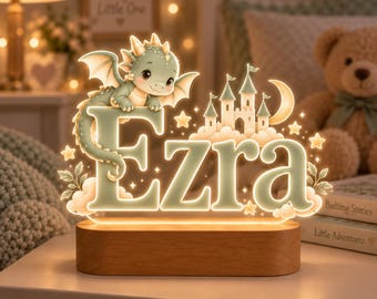 May include: A custom LED night light featuring the name Ezra in large, illuminated letters. A small green dragon sits atop the letter E, next to a castle, a crescent moon, and stars. The acrylic design rests on a smooth, rectangular wooden base.