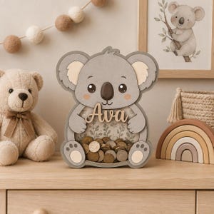Può includere: Un salvadanaio in legno a forma di koala con un pannello frontale trasparente riempito di monete. Il koala è grigio con accenti beige chiaro sulle orecchie e sui piedi. Il nome Ava è scritto in lettere di legno corsive sulla pancia.