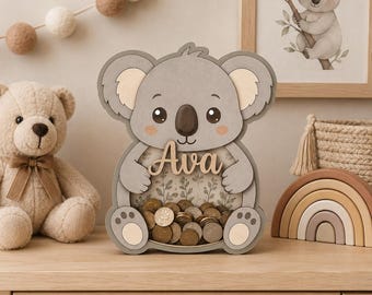 May include: A wooden koala-shaped piggy bank with a clear front panel filled with coins. The koala is gray with light beige accents on the ears and feet. The name Ava is written in wooden script letters across the center of the koala's belly.
