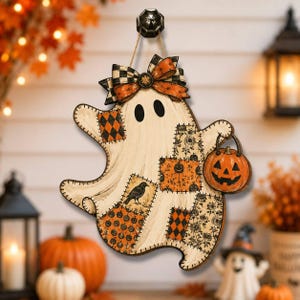 May include: A hanging ghost decoration featuring a cream-coloured body with various patchwork-style patterns including black crows, orange pumpkins, and black and orange diamonds. The ghost wears an orange and black plaid bow and holds a jack-o'-lantern.