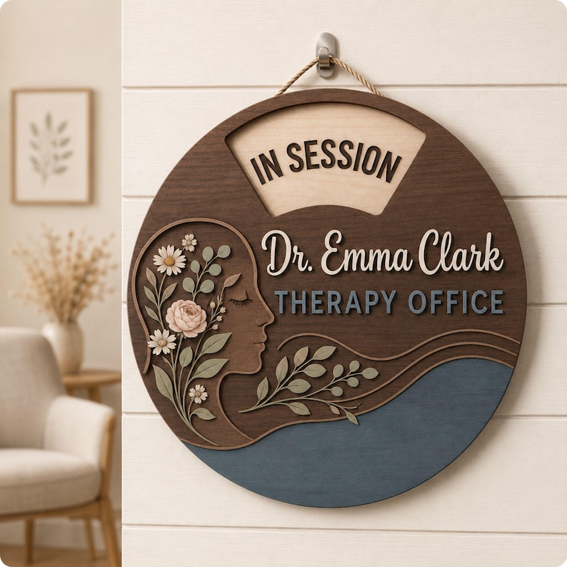 May include: A round wooden wall sign featuring the text IN SESSION, Dr. Emma Clark, and THERAPY OFFICE. The design includes a profile silhouette of a face adorned with layered floral and leaf cutouts in shades of cream, green, and blue.