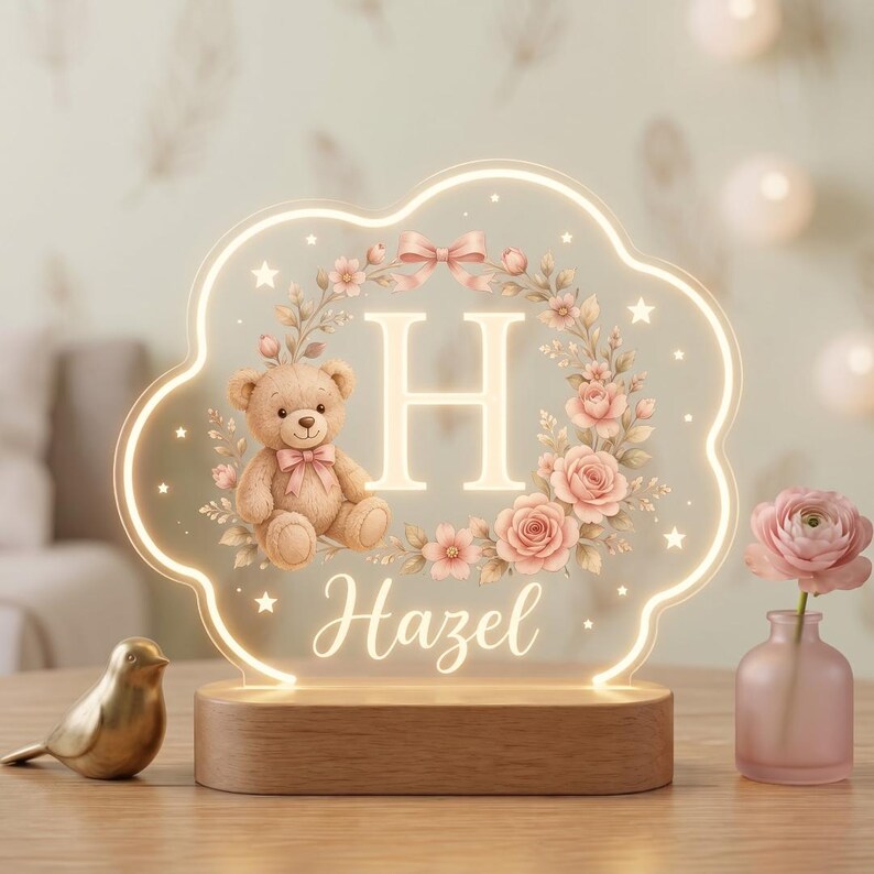 May include: A cloud-shaped acrylic night light with a wooden base featuring an illuminated letter H, a teddy bear, and a floral wreath. The design includes the name Hazel in script font, small stars, and a pink bow, all glowing with warm light.