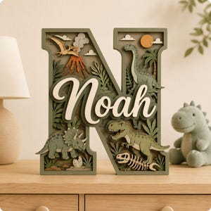 May include: A layered wooden letter N featuring a dinosaur theme with a volcano, a pterodactyl, a triceratops, a brachiosaurus, a T-Rex, and a fossil skeleton. The name Noah is written in white script across the centre of the green letter.