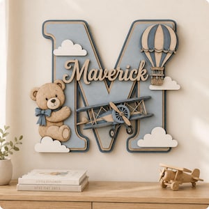 Può includere: Una grande lettera M decorativa in legno color azzurro e beige, con il nome Maverick in corsivo. È decorata con un orsacchiotto beige con un fiocco blu, un biplano blu, una mongolfiera blu e beige e diverse nuvole di colore bianco.
