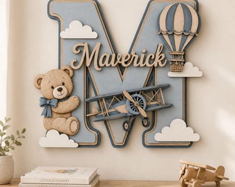 May include: A large wooden letter M wall decoration in light blue and tan, featuring the name Maverick in script. It is adorned with a tan teddy bear wearing a blue bow, a blue biplane, a blue and tan hot air balloon, and white clouds.
