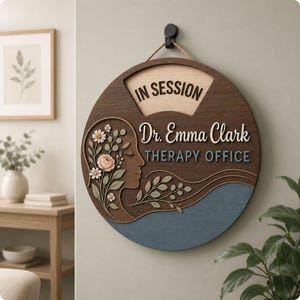 May include: A circular wooden wall sign featuring the text IN SESSION, Dr. Emma Clark, and THERAPY OFFICE. The design includes a profile silhouette of a face adorned with layered floral and leaf motifs in shades of pink, green, and blue.