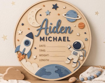 Puede incluir: Una placa de pared circular de madera con temática espacial que incluye un cohete, un astronauta, una luna y planetas. El texto dice Aiden MICHAEL, seguido de líneas para DATE, TIME, WEIGHT y LENGTH. Tonos de azul, crema y naranja.