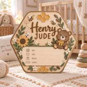 Könnte beinhalten: Ein sechseckiges hölzernes Geburtsankündigungsschild mit dem Namen Henry Jude in dunkelbrauner Schrift. Das Schild ist mit einem braunen Bären mit Honigtopf, zwei Bienen, einer Sonnenblume, grünen Blättern, weißen Blumen und Waben-Symbolen verziert.