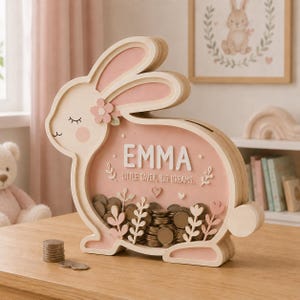 Può includere: Un salvadanaio in legno a forma di coniglio con una cornice in legno chiaro e un pannello frontale in acrilico rosa. Il nome EMMA è stampato in bianco, seguito dal testo LITTLE SAVER, BIG DREAMS. L'interno è riempito di monete argentate.