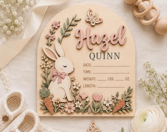 Peut inclure: Une plaque de naissance en bois en forme d'arche avec des découpes 3D représentant un lapin avec un nœud rose, des fleurs, des carottes et des papillons. Le texte indique Hazel QUINN avec des lignes pour DATE, TIME, WEIGHT, LBS, OZ et LENGTH.