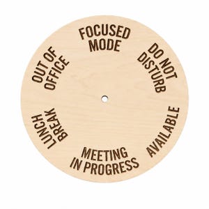 May include: A circular light-colored wooden status indicator sign featuring a central hole and laser-engraved text. The text options arranged around the center are FOCUSED MODE, DO NOT DISTURB, AVAILABLE, MEETING IN PROGRESS, LUNCH BREAK, and OUT OF OFFICE.