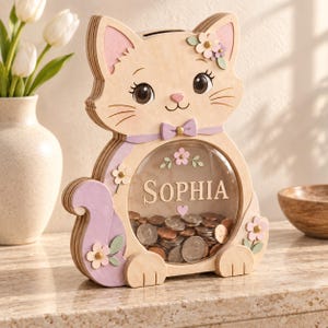 Könnte beinhalten: Eine hölzerne Spardose in Katzenform mit einem runden Sichtfenster, das den Namen Sophia und eine Sammlung von Münzen im Inneren zeigt. Die Katze hat helle Holztöne, lavendelfarbene Akzente und kleine Blumendekorationen an Ohren und Körper.