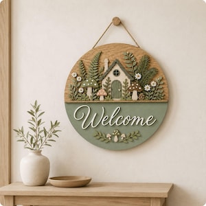 May include: A circular wooden wall sign featuring a layered 3D design of a small house, ferns, mushrooms, and white flowers on the top half. The bottom half is painted sage green with the white script text Welcome and small mushroom accents.