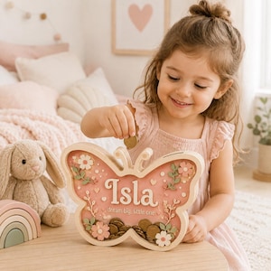 Può includere: Un salvadanaio in legno a forma di farfalla con pannello frontale trasparente, che riporta il nome Isla e la scritta dream big, little one. Il design presenta decorazioni floreali rosa e bianche, con monete dorate visibili alla base.
