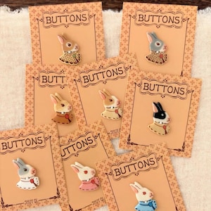 May include: Eight individual rabbit-shaped buttons, each mounted on a tan card labeled BUTTONS and SHUY QI. The buttons feature rabbits in various colors including white, grey, brown, and black, wearing different colored collars and outfits.