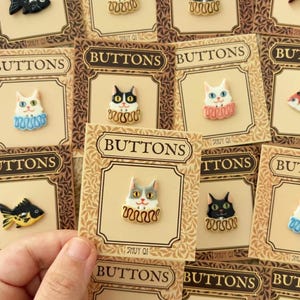 May include: A collection of individual buttons mounted on tan cards labeled BUTTONS and SHUY QI. Each button features a unique ceramic design, including various cats with colorful ruffled collars and black or gold fish shapes on a light background.
