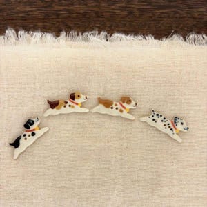 May include: Four ceramic dog-shaped buttons in a running pose. One is black and white, two are brown and white with spots, and one is white with black spots. Each dog wears a small orange collar with a yellow tag, featuring a smooth, glossy finish.
