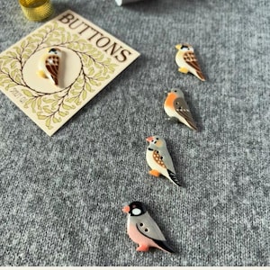 May include: Four ceramic bird-shaped buttons with detailed markings in shades of brown, gray, white, and orange, arranged near a cream-colored card featuring a wreath illustration and the word BUTTONS printed in black capital letters.