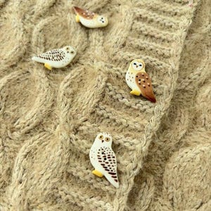 May include: Four owl-shaped buttons with detailed brown and white plumage patterns are attached to a beige cable-knit fabric. Each button features a distinct owl design with small yellow feet, showcasing a variety of poses and markings on the surface.