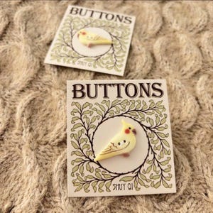 May include: Two square white cards labeled BUTTONS and SHUY QI, each featuring a light yellow cockatiel bird button with orange cheek patches and brown wing details, centered within a decorative green leaf wreath illustration on the card.