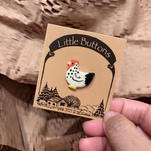 May include: A ceramic button shaped like a chicken with a white body, black spots, a red comb, and a yellow beak, attached to a tan display card labeled Little Buttons with a black ink illustration of a house and trees at the bottom.