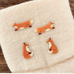 May include: Four fox-shaped buttons featuring a smooth orange and white finish. Two buttons depict foxes in a horizontal running pose, while the other two buttons show foxes in a seated position. Each button includes two small holes for attachment.