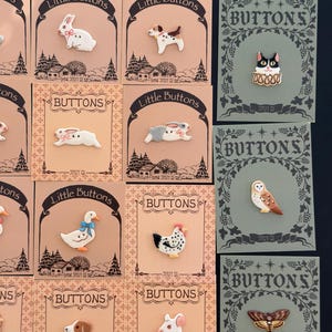Peut inclure: Collection de boutons décoratifs avec des motifs d'animaux, dont des lapins, des chiens, des chats, des hiboux et des papillons. Chaque bouton est monté sur une carte avec le texte "Little Buttons" ou "BUTTONS". Les boutons sont colorés et détaillés.
