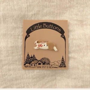 Puede incluir: Un botón con forma de conejo corriendo con coloración marrón y blanca y un pequeño detalle de lazo rojo. El botón está sujeto a una tarjeta de presentación color canela con el texto Little Buttons y una ilustración gráfica negra de una casa.