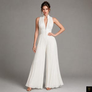 Peut inclure: Une combinaison blanche sans manches avec un col montant, une fermeture éclair verticale sur le devant et un corsage ajusté se prolongeant par un pantalon à jambes larges. Le vêtement est entièrement fait d'un tissu finement plissé.