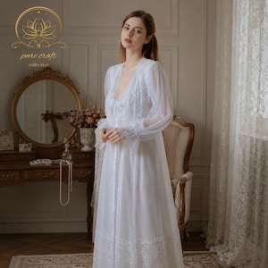 Puede incluir: Un camisón blanco de manga larga con detalles de encaje. El camisón presenta un escote en V y mangas transparentes. La imagen incluye un logotipo dorado con el texto "pure craft collection".