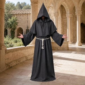 May include: A floor-length black robe featuring a pointed hood, wide sleeves, and a shoulder capelet. The garment is cinched at the waist with a thick, off-white knotted rope belt that hangs down the front, creating a structured, full-length silhouette.