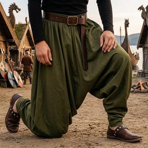 May include: An olive green, loose-fitting pants with a brown leather belt and shoes. The person is kneeling in front of a Viking village setting. The pants are gathered at the ankles. The background includes wooden buildings, shields, and axes.
