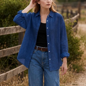 May include: A long-sleeved, button-down shirt in a deep blue colour, worn over a black vest top and paired with blue denim jeans featuring a brown leather belt. The shirt has a classic collar, white buttons, and cuffs that are rolled up at the wrists.